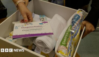A white cardboard box with the baby bundle items, including clothing, blankets, bibs, muslin cloths, a thermometer, a bilingual book and a playmat. On top is a note which reads 'congratulations...on your bundle of joy', which a hand with pink nail polish reaching in the box to pick it up.