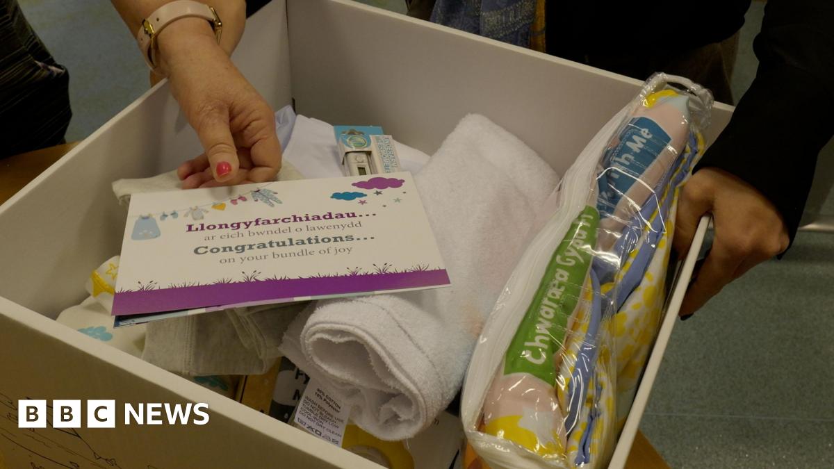 A white cardboard box with the baby bundle items, including clothing, blankets, bibs, muslin cloths, a thermometer, a bilingual book and a playmat. On top is a note which reads 'congratulations...on your bundle of joy', which a hand with pink nail polish reaching in the box to pick it up.