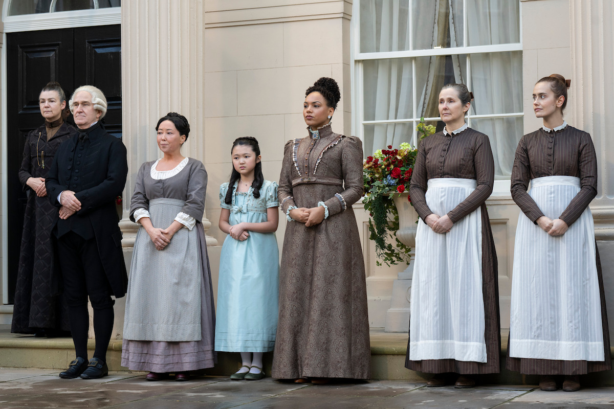 A group of people in historical clothing stand outside a building with large pillars and windows, including women in long dresses and aprons, a man in black, and a young girl, all in a formal, staged pose.