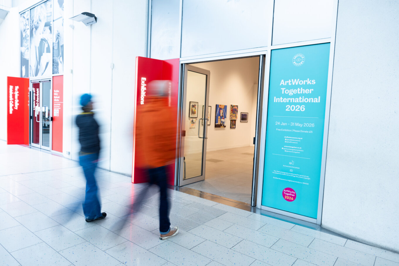 A photo of two people, slightly blurred, walking past the entrance to an exhibition called "ArtWorks Together" in an art gallery