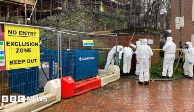 A group of police forensic investigators carrying poles, wearing white boiler suits. They are looking at a green bin. Next to them is temporary fencing with a yellow sign on it saying 'No entry, exclusion zone, keep out'