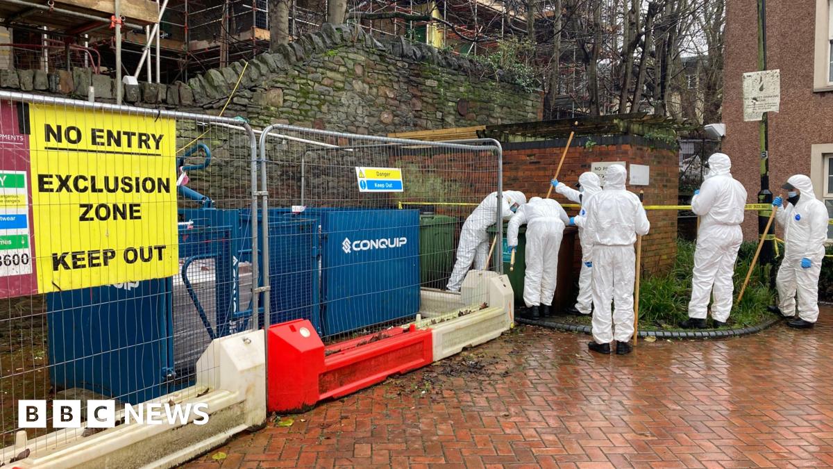 A group of police forensic investigators carrying poles, wearing white boiler suits. They are looking at a green bin. Next to them is temporary fencing with a yellow sign on it saying 'No entry, exclusion zone, keep out'