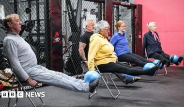 Five members of the class four older and one younger, sit on chairs with their legs outstretched balancing blue balls on their feet