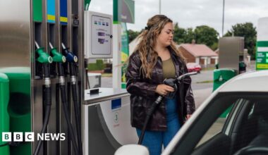 Young woman with long dark blonde hair, wearing black leather jacket with khaki green top and dark blue jeans fills her car with fuel at a forecourt
