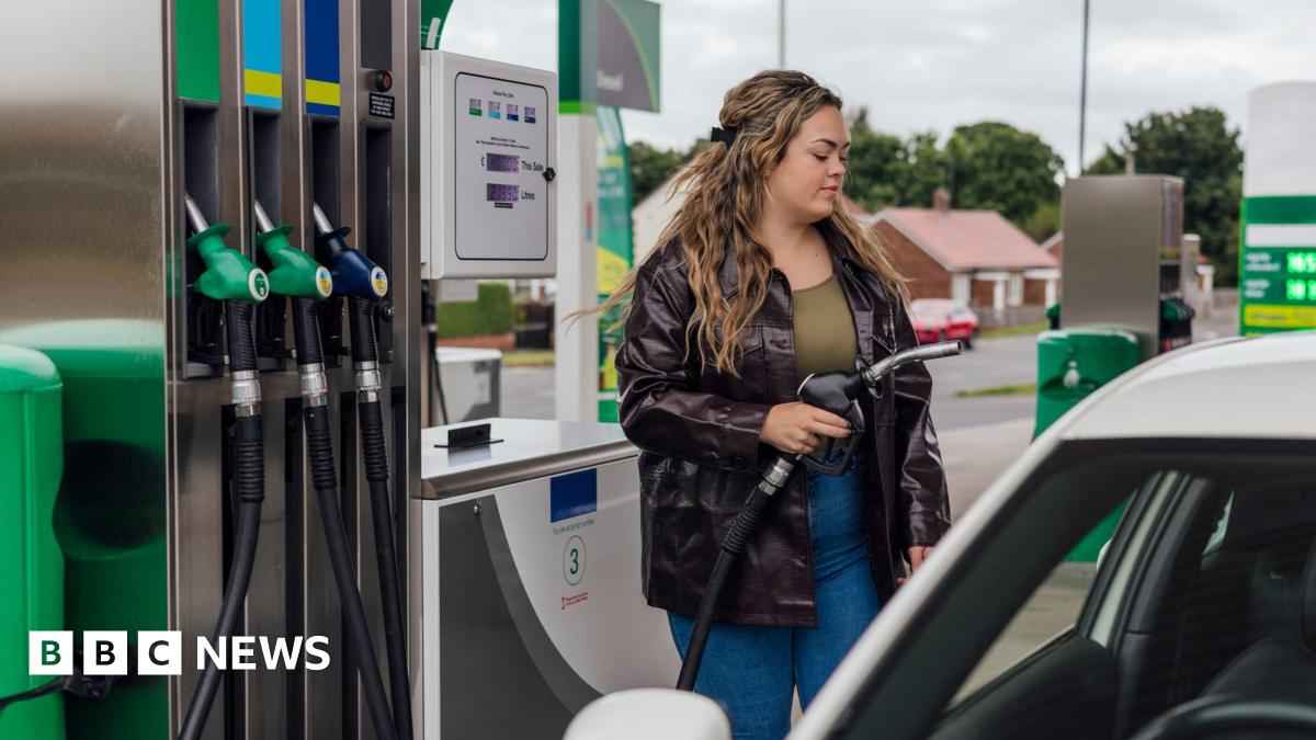 Young woman with long dark blonde hair, wearing black leather jacket with khaki green top and dark blue jeans fills her car with fuel at a forecourt