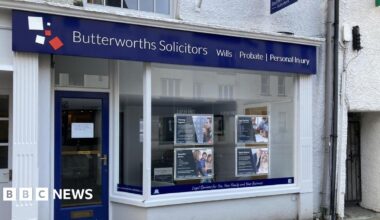 The front of a solicitors office. There is a big glass window with some advertising panels hung in it. There is a blue sign above the whole shop frontage which says 'Butterworths Solicitors.' The door is glass with a blue trim. In the centre of the door is a white piece of paper stuck to it with writing on it.