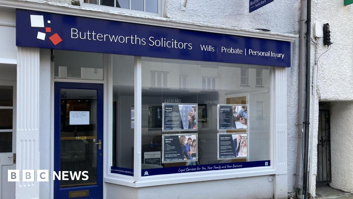 The front of a solicitors office. There is a big glass window with some advertising panels hung in it. There is a blue sign above the whole shop frontage which says 'Butterworths Solicitors.' The door is glass with a blue trim. In the centre of the door is a white piece of paper stuck to it with writing on it.