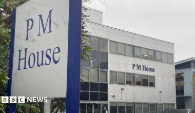 A large office building with the name “PM House” displayed prominently on its exterior. The building has a modern, rectangular design with multiple rows of windows and light grey cladding. In the foreground, a tall sign also reads “PM House” in blue lettering on a white background, framed by a dark blue border. Several cars are parked in front of the building, suggesting a business or office complex.
