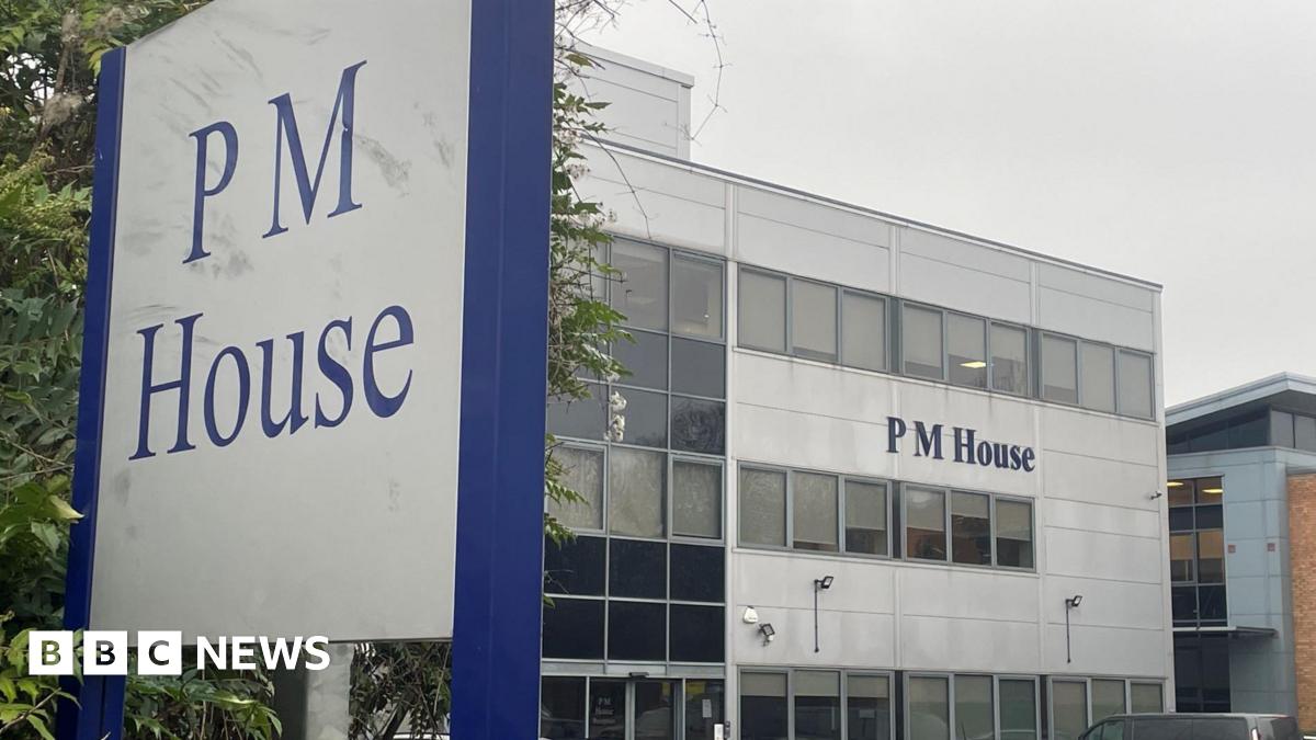A large office building with the name “PM House” displayed prominently on its exterior. The building has a modern, rectangular design with multiple rows of windows and light grey cladding. In the foreground, a tall sign also reads “PM House” in blue lettering on a white background, framed by a dark blue border. Several cars are parked in front of the building, suggesting a business or office complex.