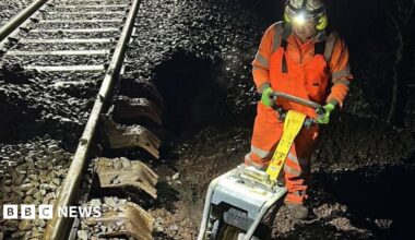 A rail engineer works at night on repairs to a line in Devon. He is wearing orange high-vis overalls and has a light on his helmet. He also has a piece of equipment in his hands.