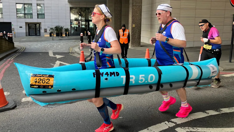 Two runners wearing sailor hats navigate the race course while carrying a large, blue inflatable boat prop around their waists.