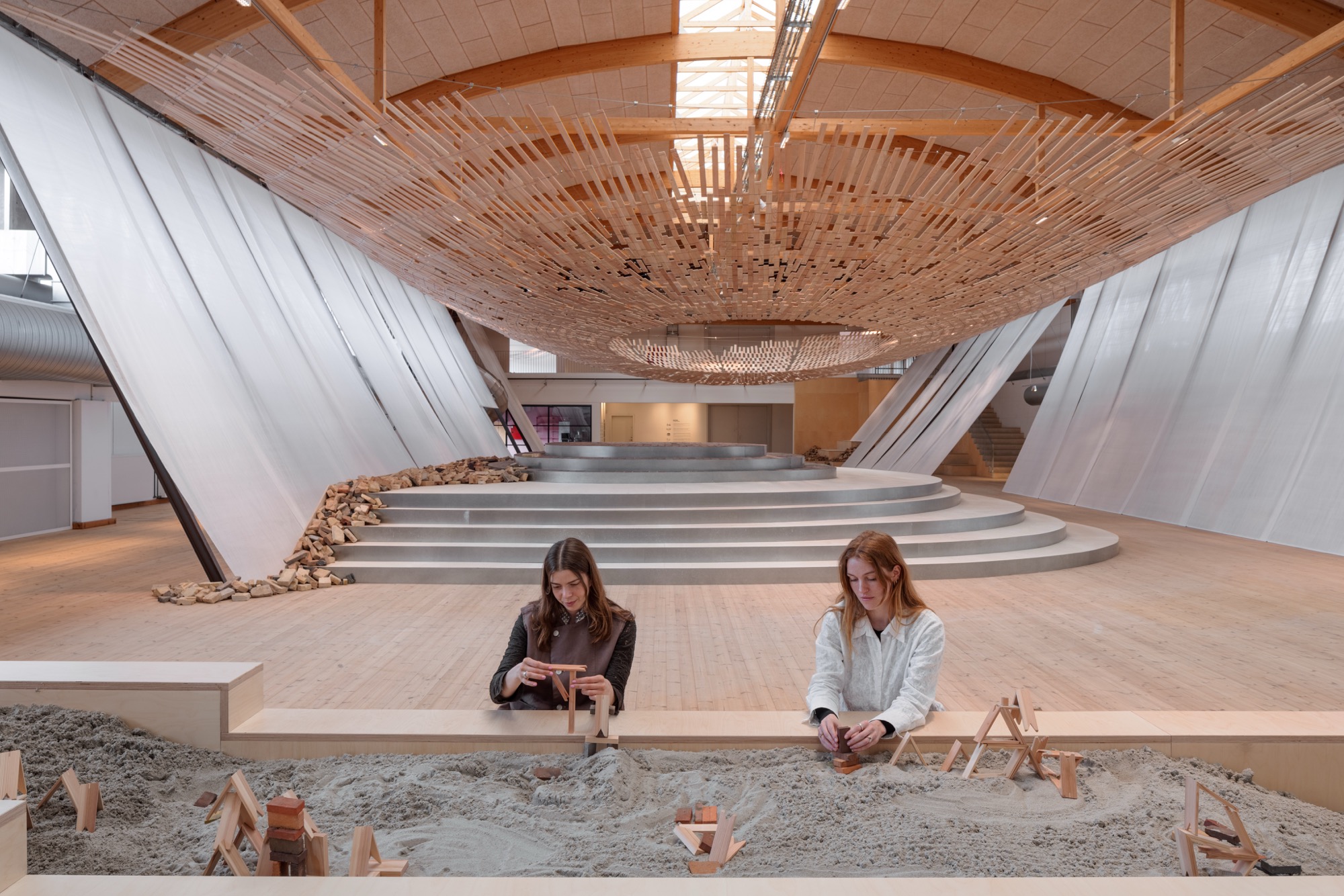 visitors work at a table and enjoy an installation in which light streams through a wooden canopy by Kengo Kuma