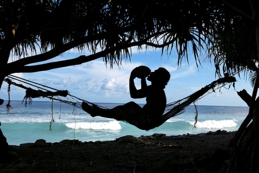 A young man drinks coconut milk while relaxing in a hammock on the shores of Funafuti.