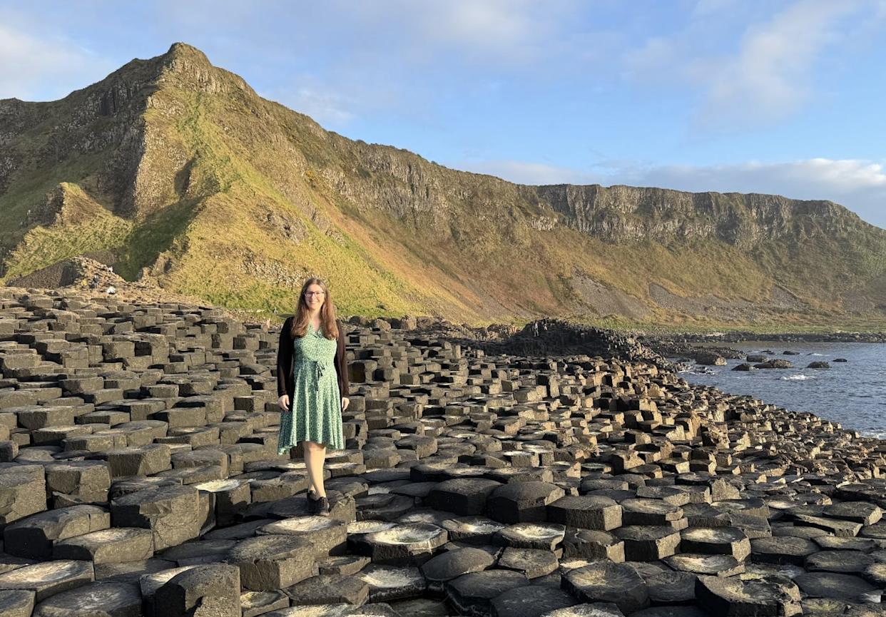 The writer standing on pillars of hardened lava at the Giant's Causeway, with hills in the background.