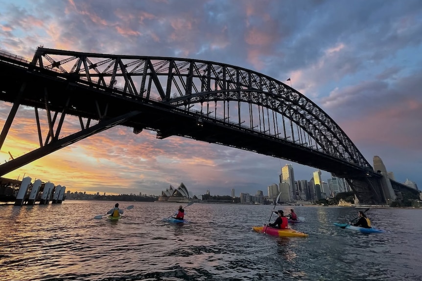 A wide landscape shot at sunrise of five kayakers under the Sydney Harbour Bridge with the Opera House and clouds backdrop.