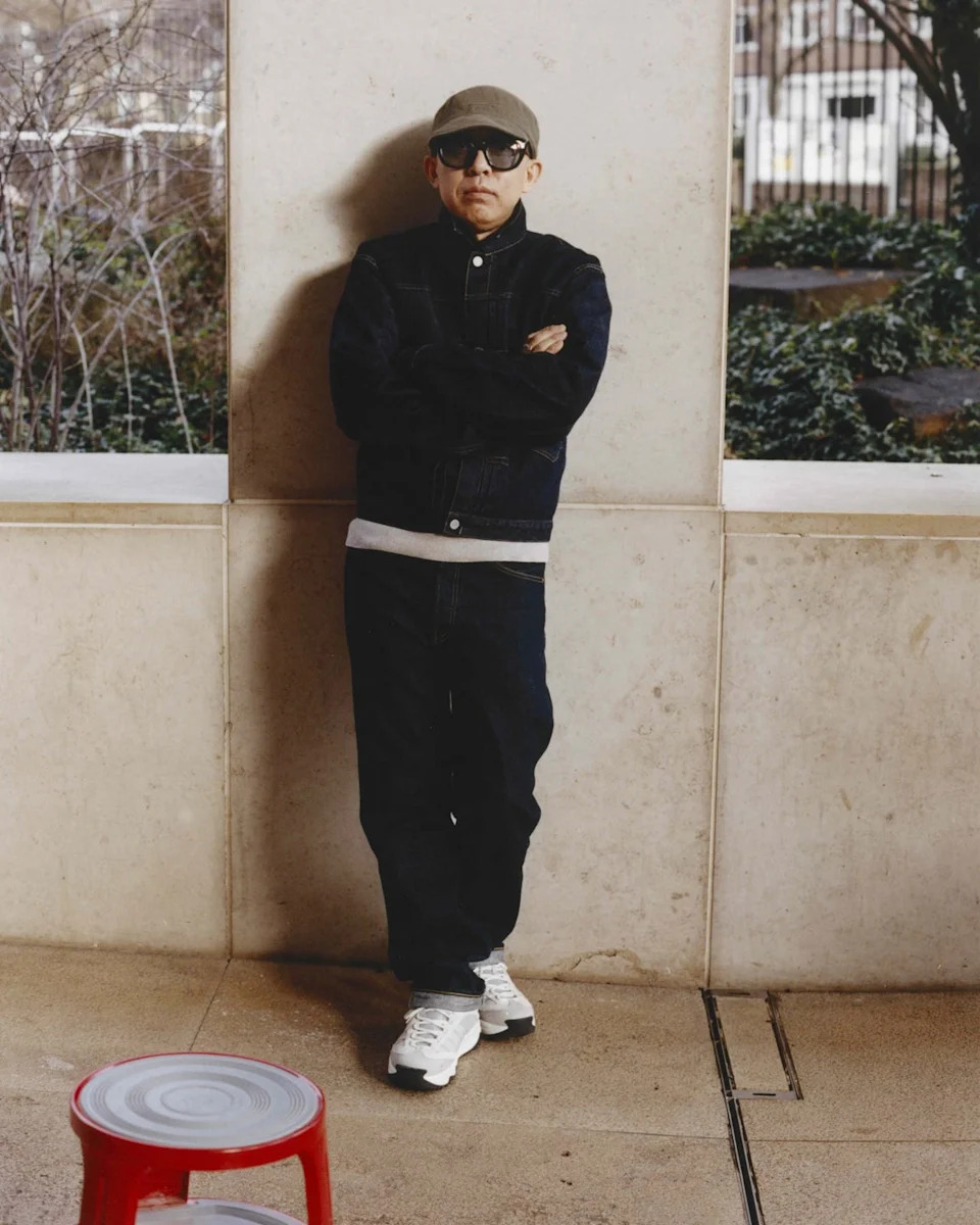 a person standing with arms crossed against a wall with a small red stool in the foreground