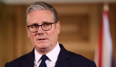 A medium close-up of Keir Starmer wearing a dark suit, polka-dot tie, and glasses, appearing to speak at a podium against a blurred wooden background and a flag.