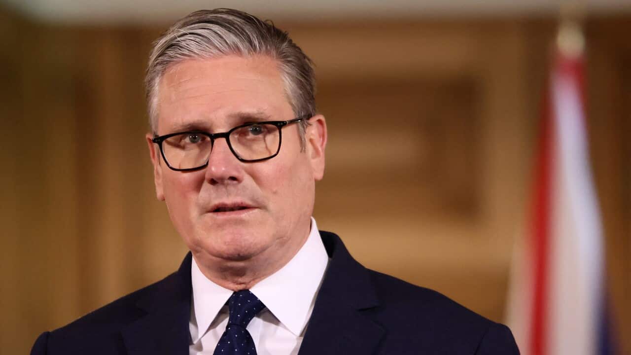 A medium close-up of Keir Starmer wearing a dark suit, polka-dot tie, and glasses, appearing to speak at a podium against a blurred wooden background and a flag.