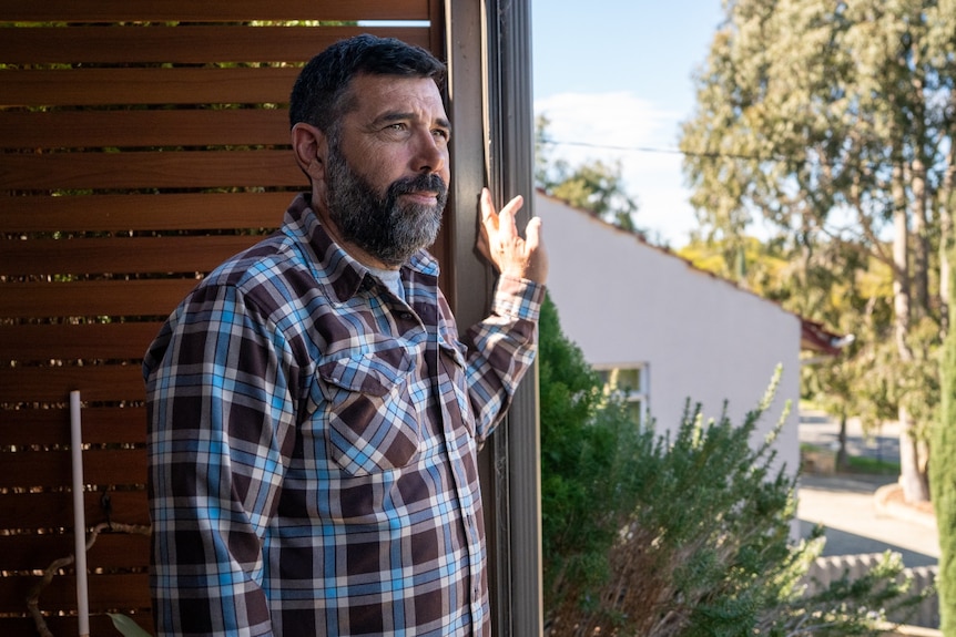 A man in a brown and blue checked shirt standing at the entrance to a house.