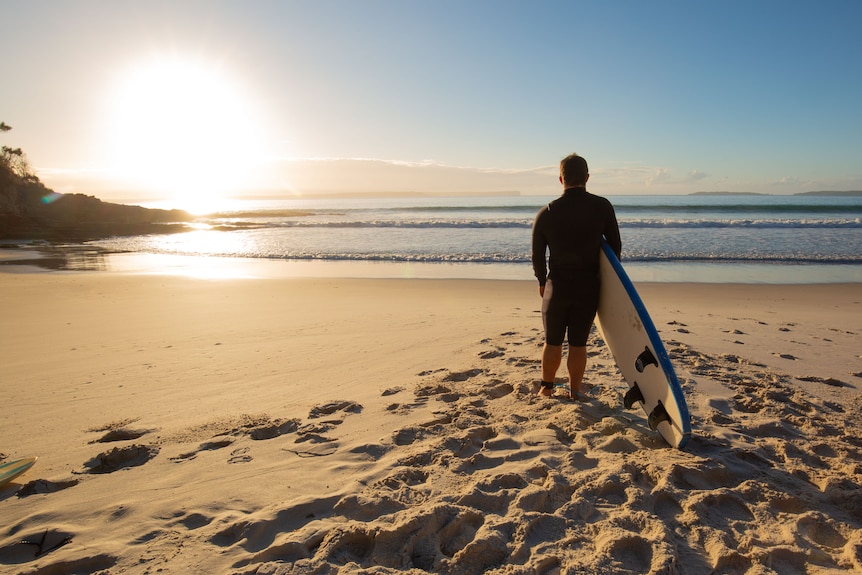 A young man stands holding a surfboard as the sun rises over the ocean