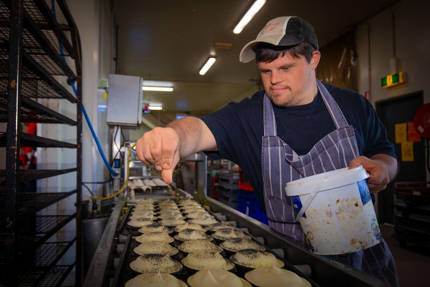 A young man wearing an apron sprinkles toppings on trays of pies