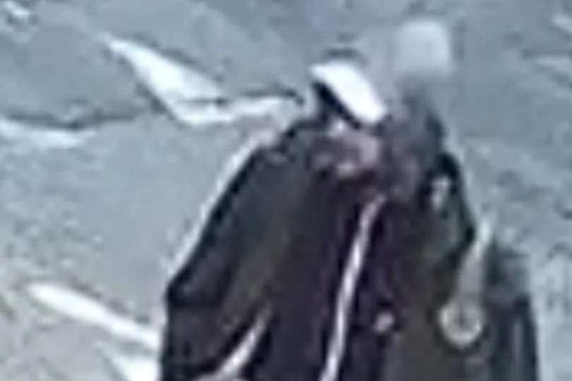 A blurry CCTV image of a white man with a beard., pictured wearing a cap, and a coat with the hood up and also wearing a darker top under the coat, light blue, stonewashed jeans and white trainers.