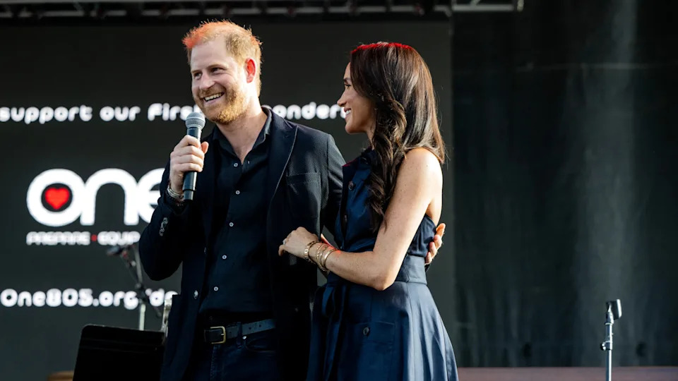 Prince Harry and Meghan Markle on stage, smiling, with Meghan holding Harry's arm. They're at an event with a microphone.