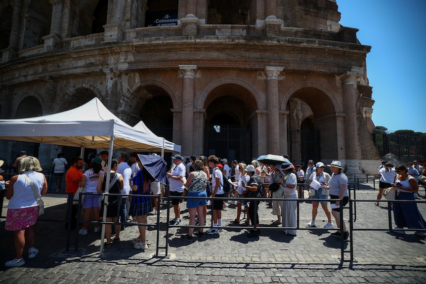 Visitors outside ancient ruins, use umbrellas for shade as they stand in line.