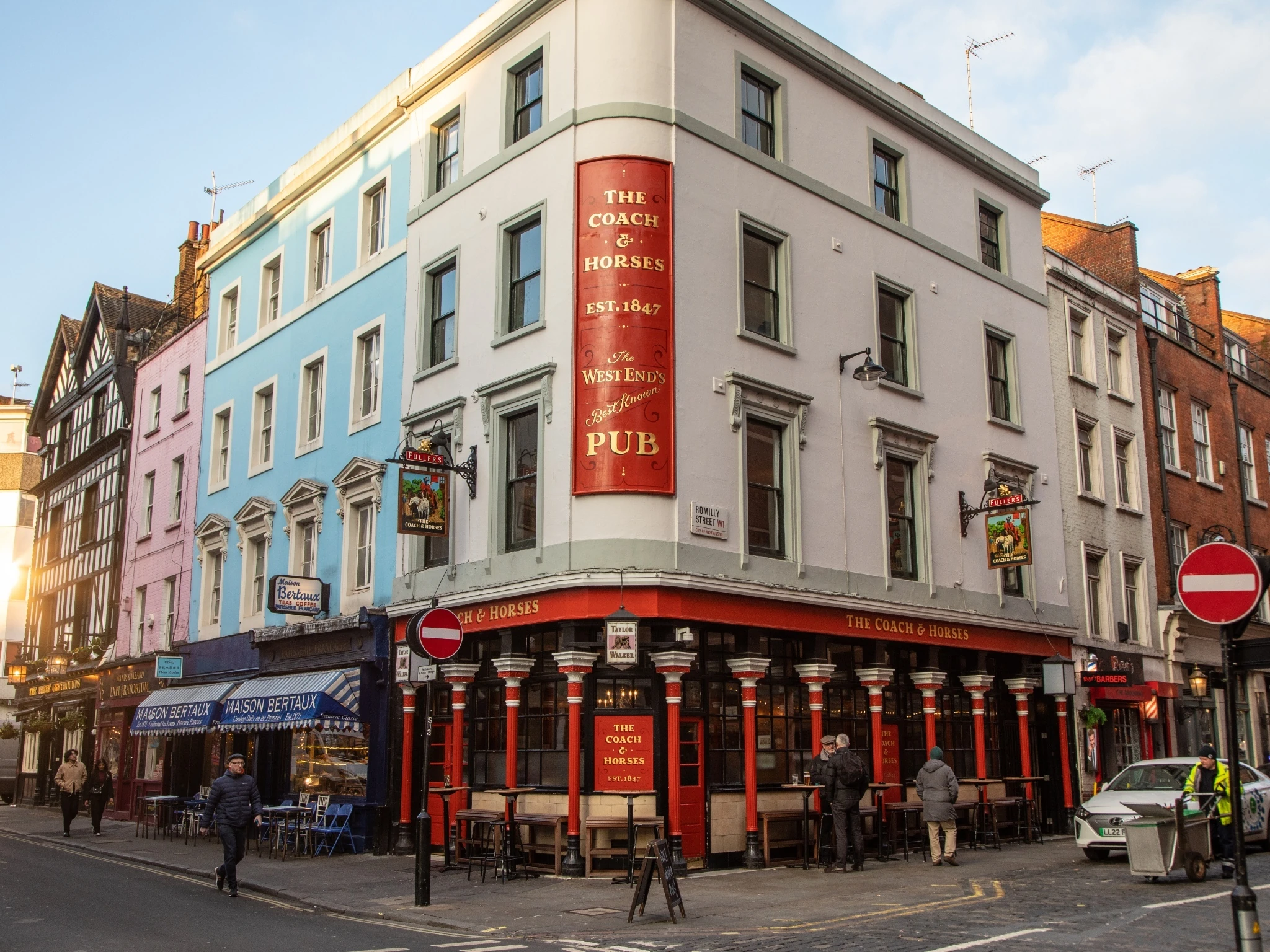 A large red sign painted with gold text hangs on the first floor level above painted red pillars decorating the exterior of the Coach & Horses, a corner pub viewed from the other side of the road