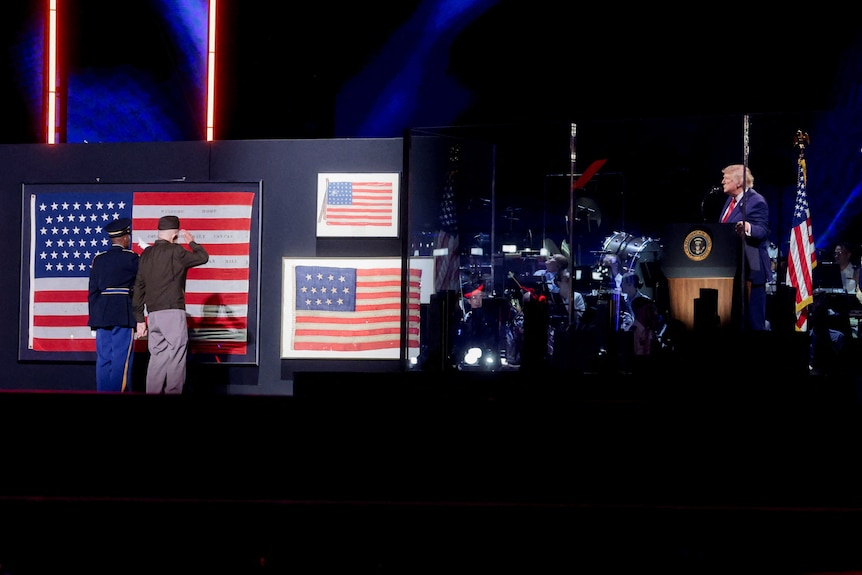 Two veteran soldiers salute to US flags on stage as Donald Trump looks on from presidential lectern