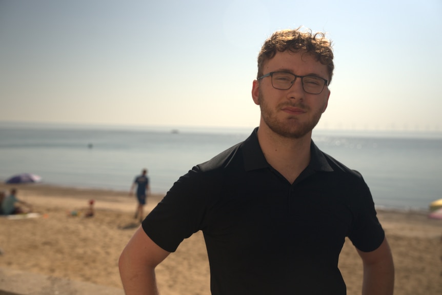 A young man with dark hair, a black t shirt and glasses stands infront of a beach landscape.