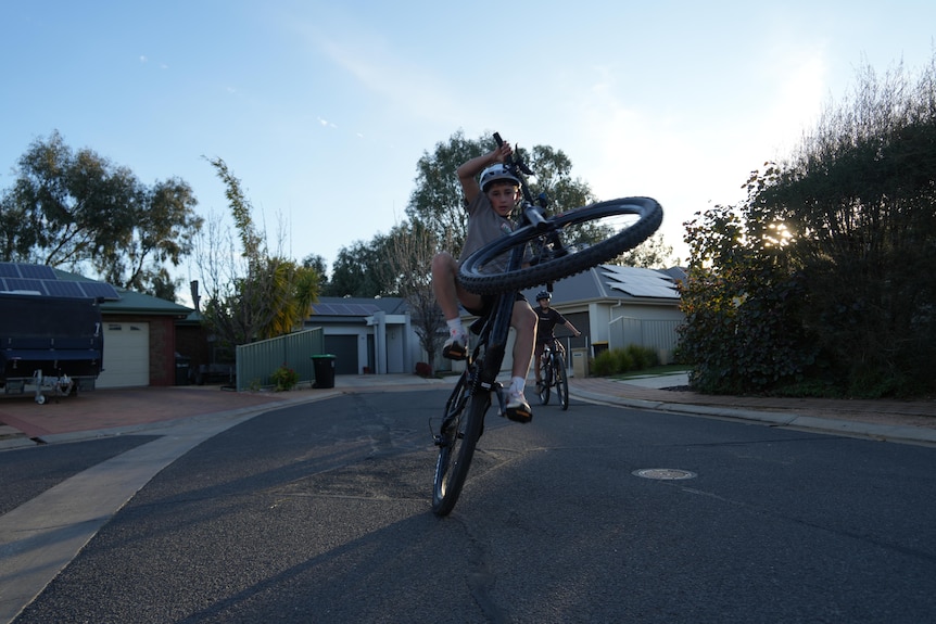 Christos performs a wheelie on his bike on a suburban street, sun casts shadow on the ground.