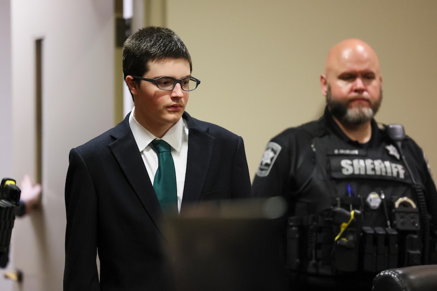 Colt Gray, in suit and tie, is led into court by a man in police uniform.