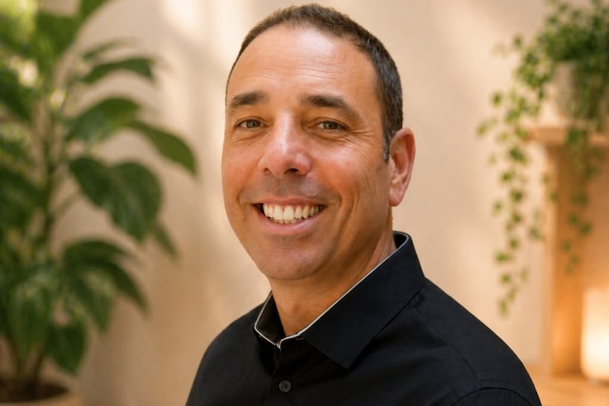 A man wearing a dark coloured shirt smiles with his mouth open. He has short dark, cropped hair and there are plants behind him.