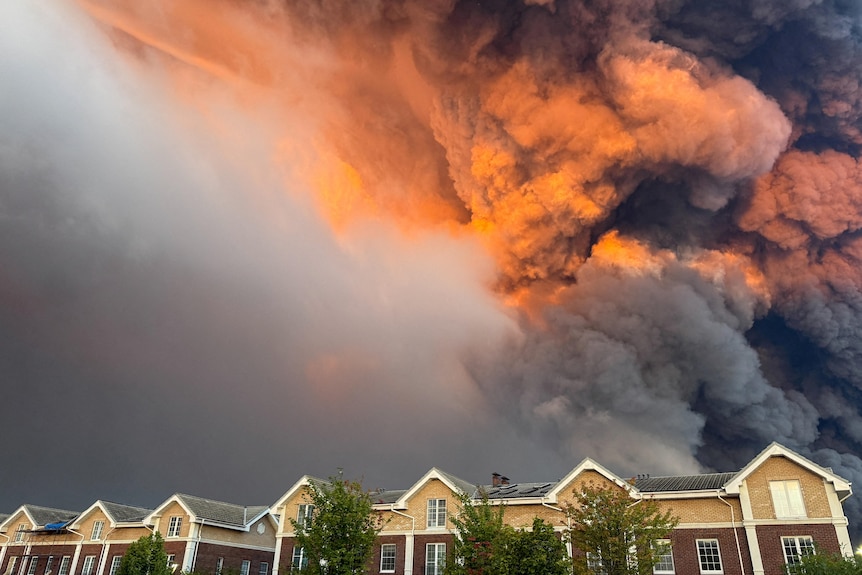A large plume of smoke reaching high into the atmosphere is seen behind a row of two-storey suburban homes.