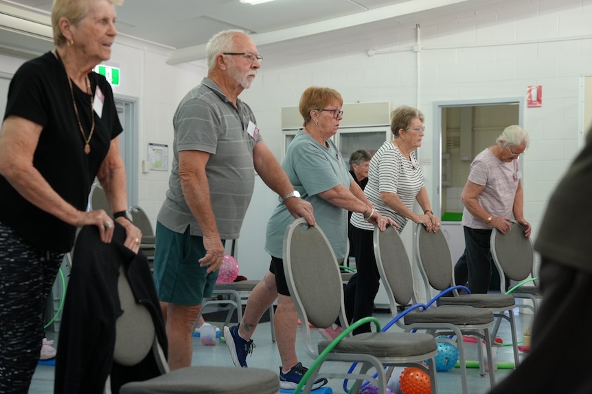 elderly men and women practicing their balance by putting one hand on a chair and raising a foot behind them