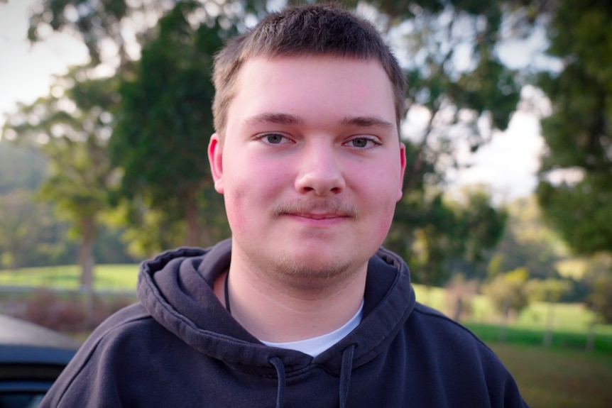 teenage boy with short, brown hair and moustache looks to camera. He wears a black hoodie.