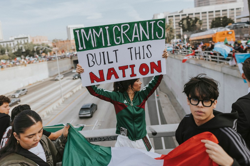 A person in a green tracksuit holding up a sign saying, “IMMIGRANTS UILT THIS NATION,” while next to a guard railing over a highway in downtown Los Angeles. In the background, cars can be seen driving on the road while crowds of protesters hold up signs and flags on both sides of the highway.
