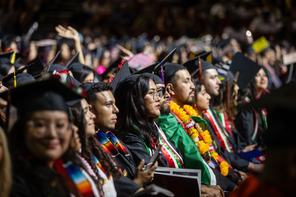 A group of college graduates sits in rows during a commencement ceremony, wearing black caps and gowns adorned with colorful stoles, leis, and sashes. The focus is on a young woman in glasses looking ahead, surrounded by classmates with celebratory decorations, including a student in a green gown and marigold leis.
