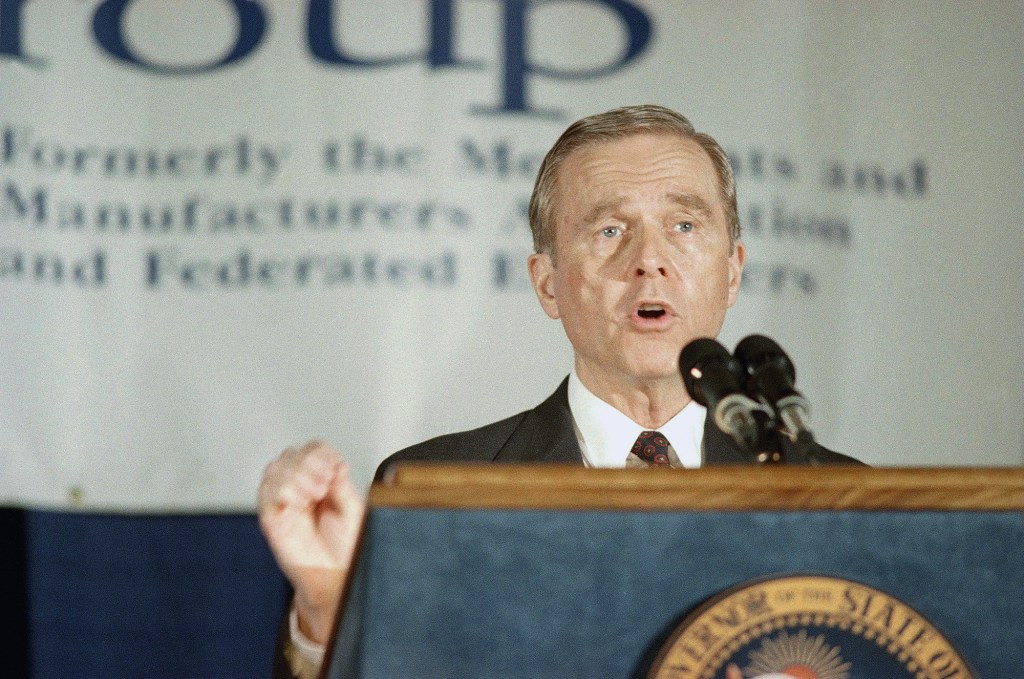 A man in a suit and tie speaks into two microphones at a podium with the seal of the State of California. He gestures with one hand raised slightly. A blurred banner in the background suggests the event is hosted by a business or trade group.