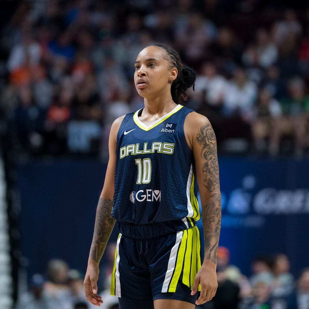 Aziaha James stands on the court in a blue Dallas Wings jersey, looking forward