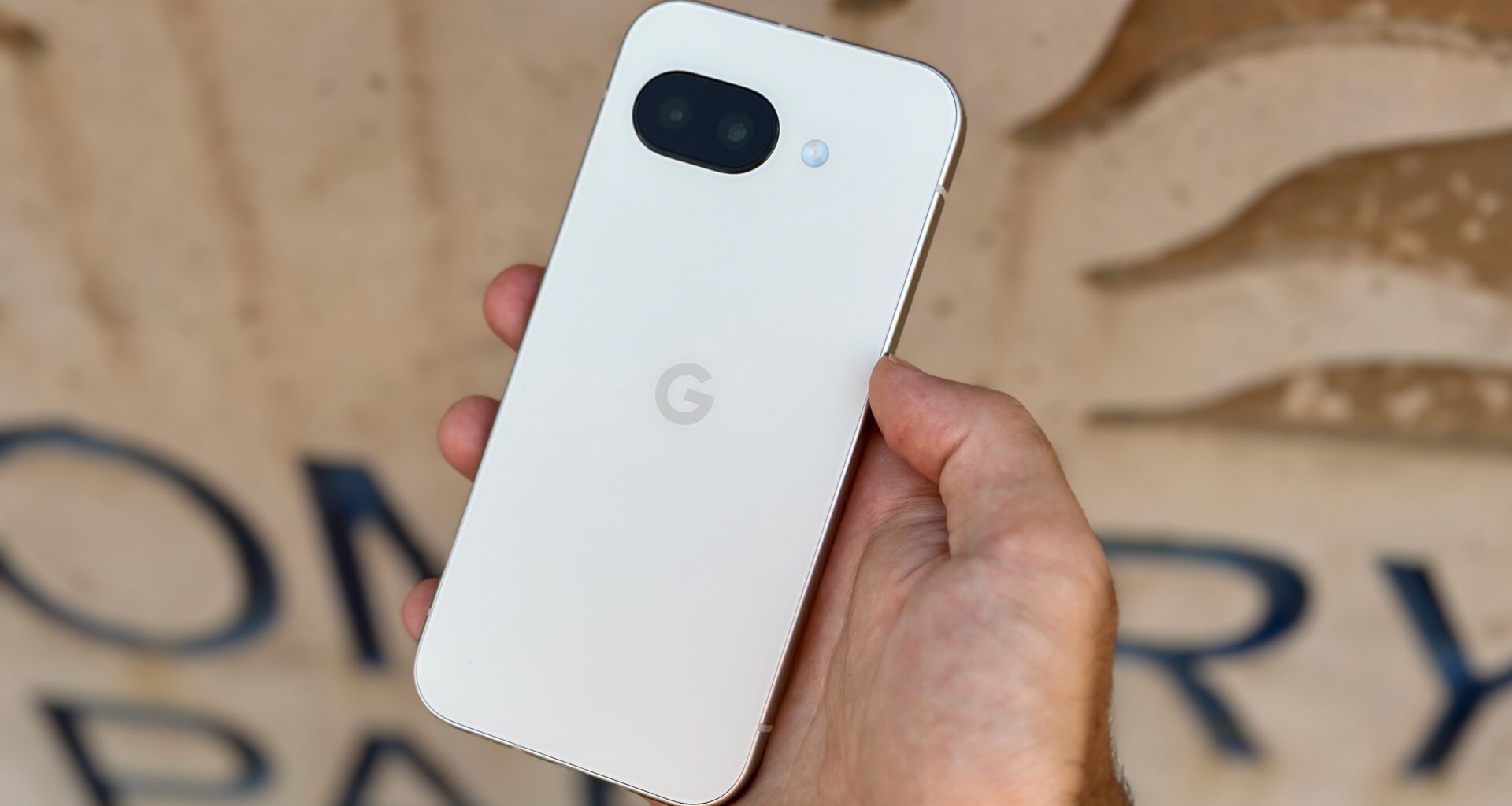 The Porcelain Google Pixel 9a held in hand in front of a tan sign.