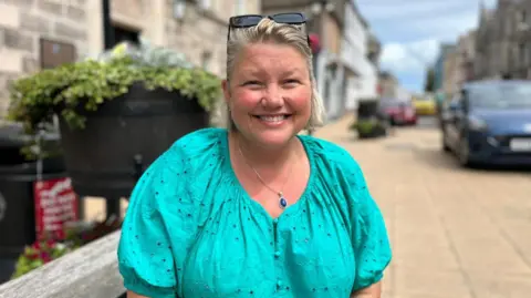 Lucy has shoulder length blonde hair and sun glasses perched on top of her head. She has a turquoise top and is smiling while she stands in Nairn High Street. 