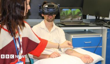 A man is sitting in a room wearing a virtual reality headset. A woman wearing two lanyards is standing next to him and is holding a metal pencil-shaped item to his forearm which is stretched out across a pillow. Behind him is a laptop showing two circles of the same photo which feature green trees and a bright blue sky. The laptop sits on the top of a desk with a monitor next to it which is off and there are blue drawers below.