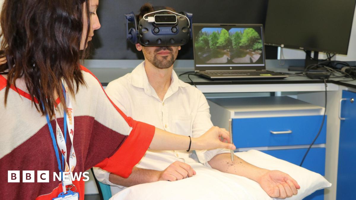 A man is sitting in a room wearing a virtual reality headset. A woman wearing two lanyards is standing next to him and is holding a metal pencil-shaped item to his forearm which is stretched out across a pillow. Behind him is a laptop showing two circles of the same photo which feature green trees and a bright blue sky. The laptop sits on the top of a desk with a monitor next to it which is off and there are blue drawers below.