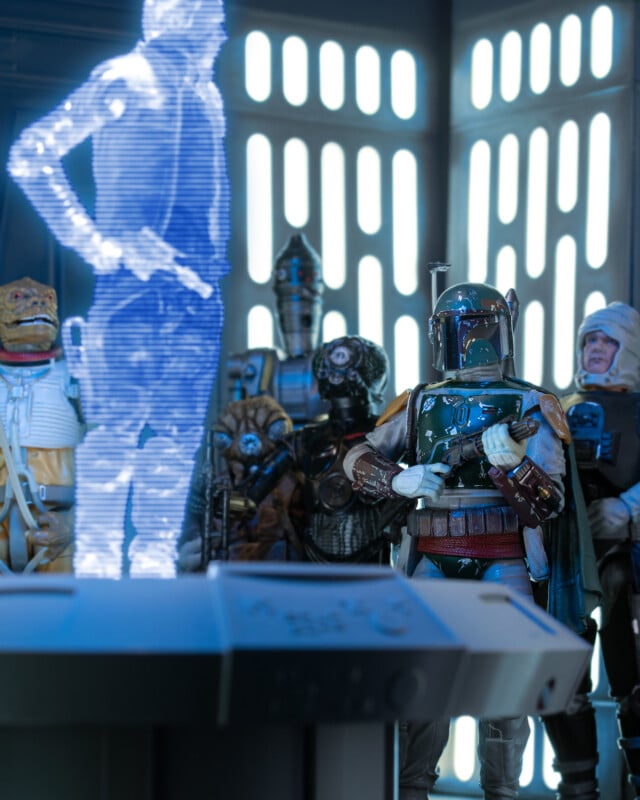 A group of Star Wars bounty hunter action figures, including Boba Fett, stand in a spaceship room, facing a blue hologram of a person projected from a console. Light streams through vertical slits in the background.