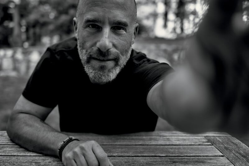 A man with a beard and short hair sits at a wooden table outdoors, reaching toward the camera. The photo is in black and white, with a blurred natural background.