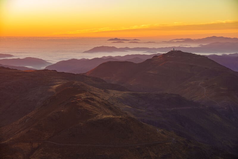 Mountain landscape at sunset with soft orange and yellow hues in the sky. Layers of mountains fade into the distance, and a white observatory sits atop one of the peaks. A winding road runs through the scene.