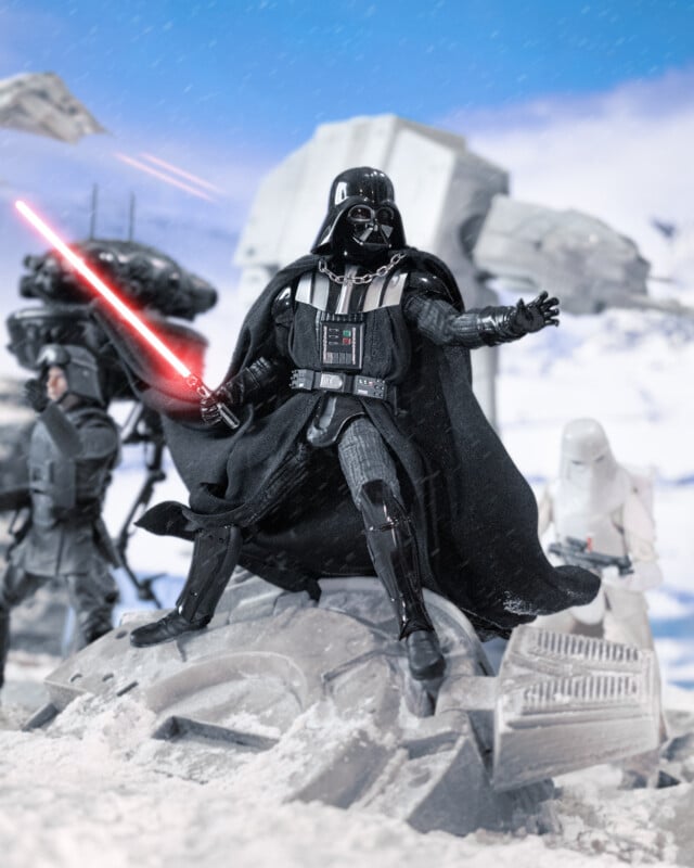 Darth Vader holding a red lightsaber stands on snowy wreckage, surrounded by Star Wars Imperial forces with AT-AT walkers and snowtroopers in the background, set on a wintry battlefield.