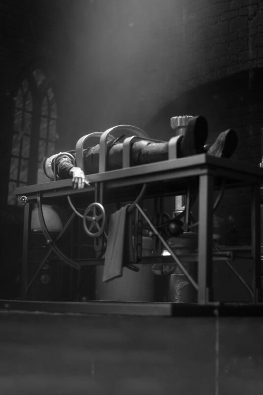 A person dressed as Frankenstein’s monster lies on a table with restraints in a dark, dramatic setting, reminiscent of a classic laboratory scene from vintage horror films.